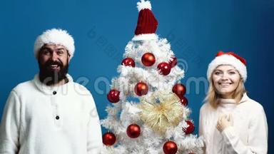 戴着<strong>新年帽</strong>子`长胡子的男人和戴着红<strong>帽</strong>子的女人站在圣诞树附近，表达情感。 概念